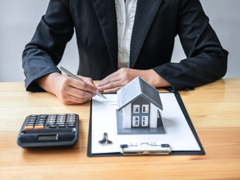 Pessoa assinando contrato com miniatura de casa e calculadora na mesa, simbolizando seguro fiança para aluguel de imóvel.