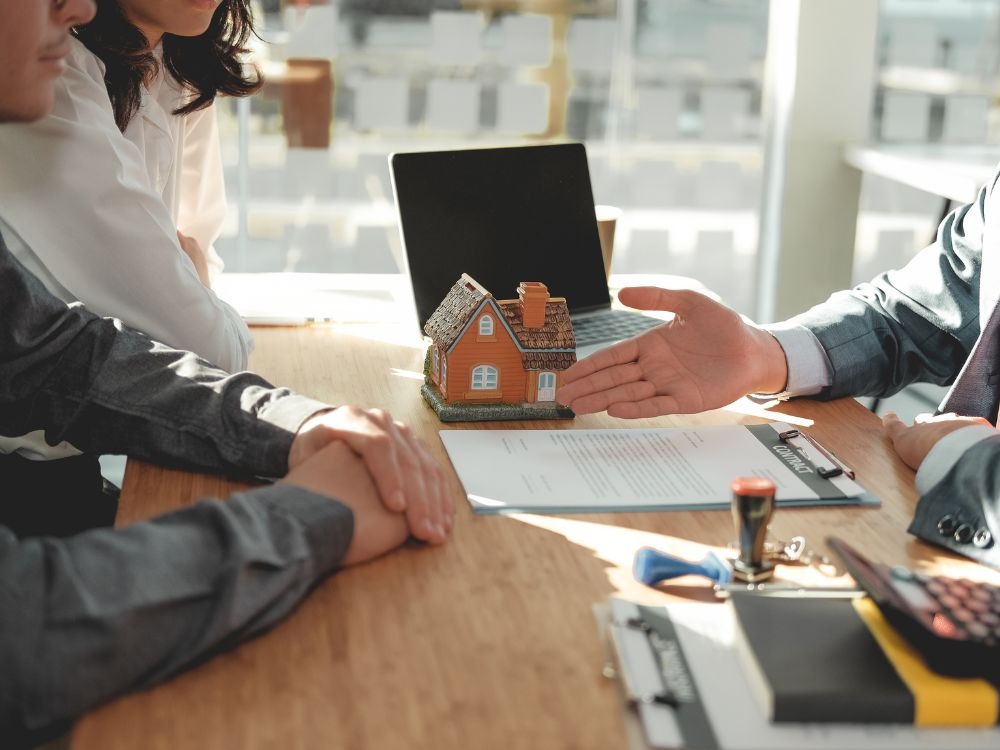 Negociação de compra e venda de imóvel com miniatura de casa e contrato em destaque sobre a mesa.