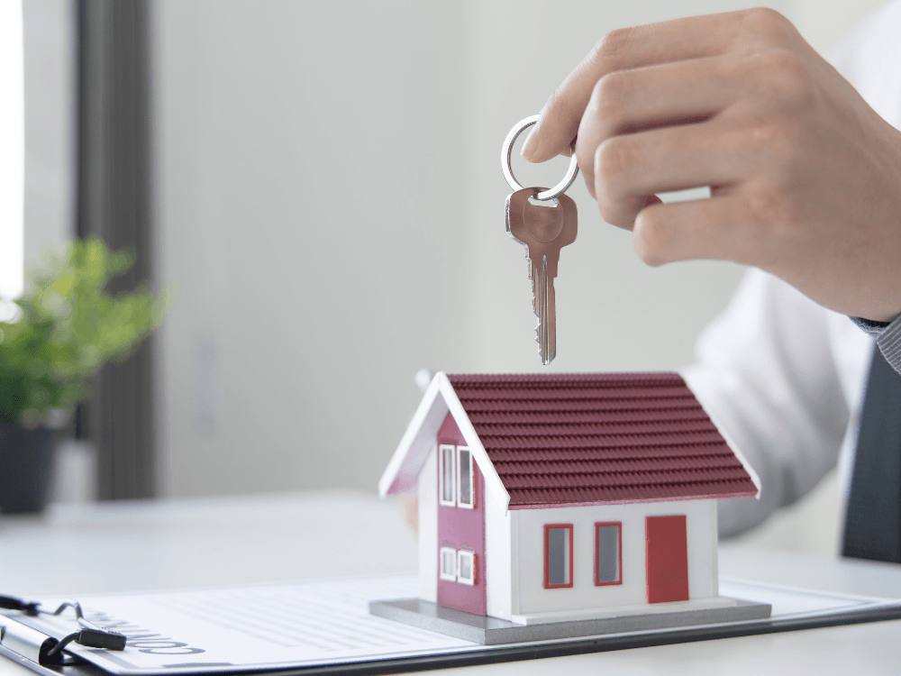Mão segurando uma chave sobre maquete de casa, ilustrando a transferência do imóvel em uma venda financiada.