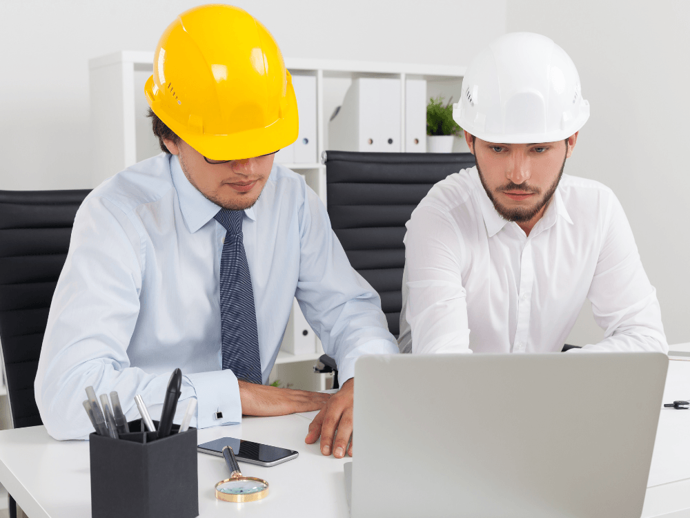 Dois profissionais com capacetes analisam informações em um notebook durante o acompanhamento de um projeto de construção civil.