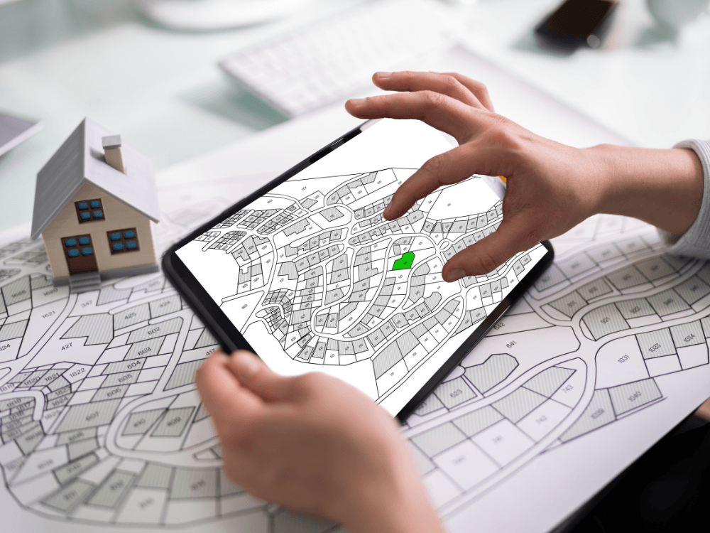 Pessoa analisando mapa cadastral em um tablet ao lado de uma maquete de casa, representando a pesquisa sobre a propriedade de um terreno.