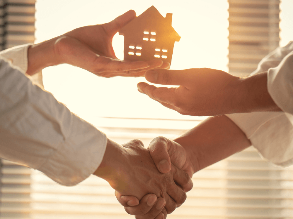 Duas pessoas apertam as mãos enquanto uma entrega uma miniatura de casa, simbolizando a negociação de um empreendimento imobiliário.