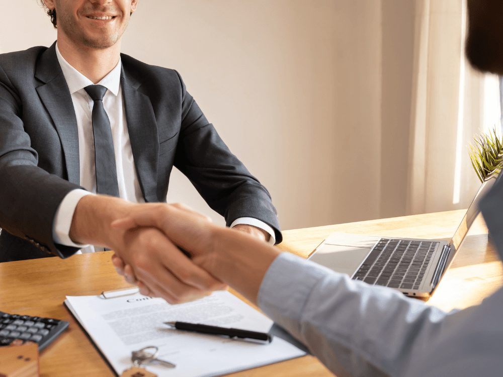 Corretor e cliente apertam as mãos durante reunião com contrato e notebook sobre a mesa, indicando negociação imobiliária.