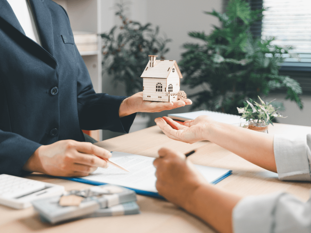 Pessoa entrega uma maquete de casa durante atendimento com documentos sobre a mesa, ilustrando a consulta aos registros imobiliários.