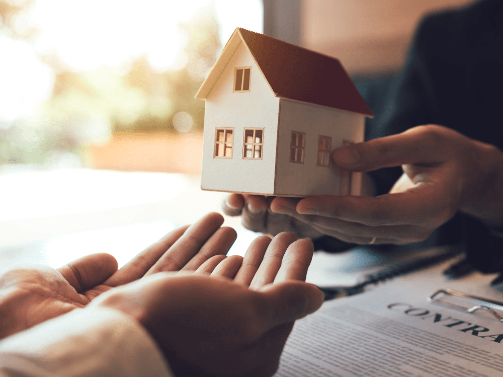 Miniatura de casa sendo entregue entre duas pessoas sobre um contrato, simbolizando a posse do imóvel após o registro imobiliário.