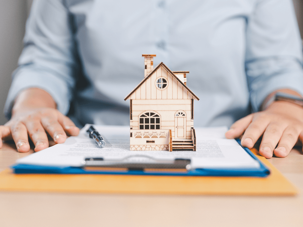 Maquete de casa posicionada sobre documentos em uma mesa, simbolizando a consulta ao histórico e aos registros de um imóvel. Tudo para explicar o que é transcrição de imóvel.