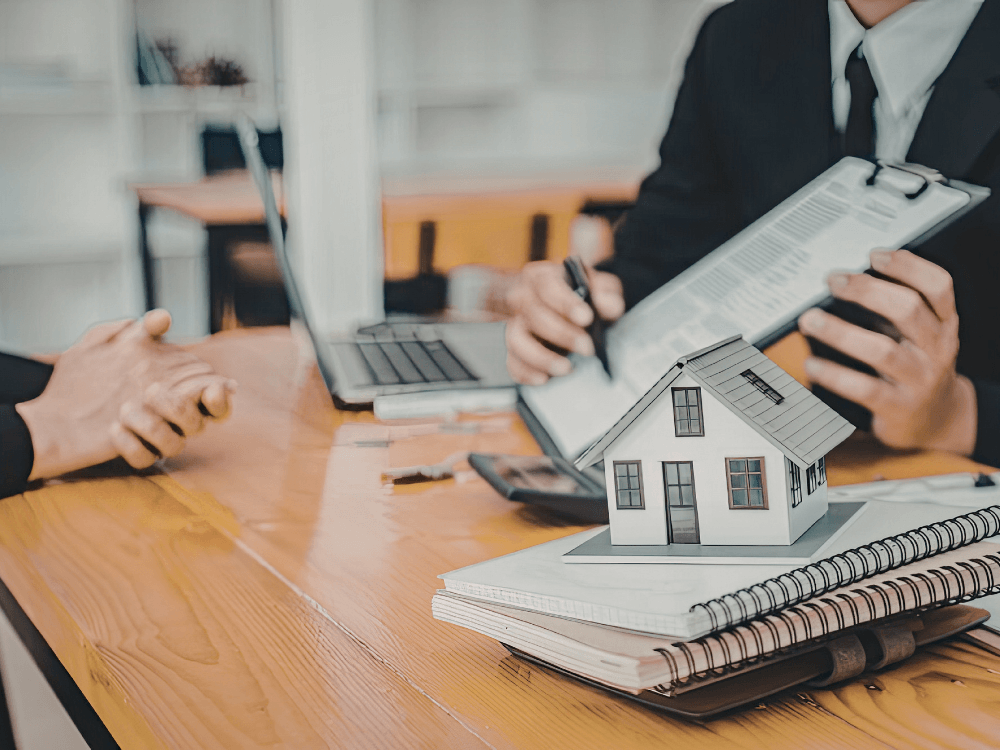 Profissional analisa documentos e contrato enquanto uma maquete de casa representa a venda de imóvel e seus custos. Tudo para explicar o que o vendedor paga na venda de imóvel.