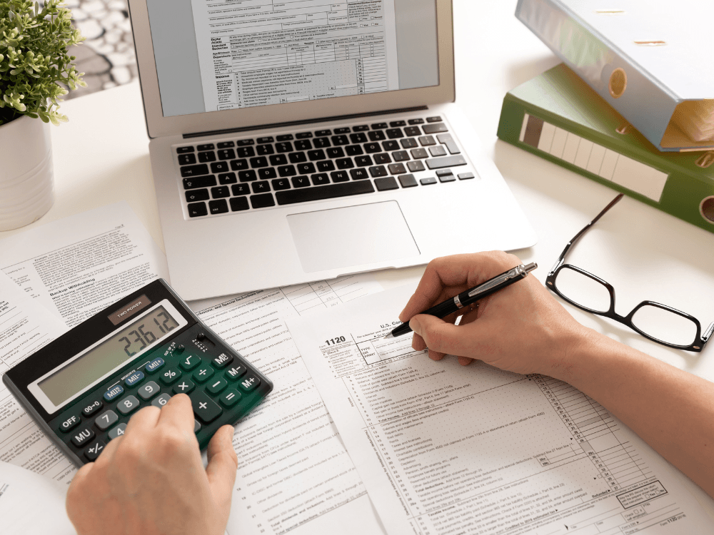 Pessoa preenche documentos e calcula valores com calculadora e computador durante a análise de despesas na venda de imóvel.
