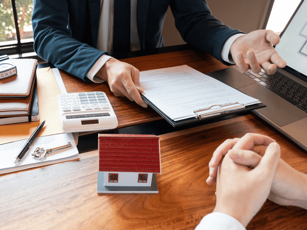 Profissional avalia contrato e cálculos financeiros com uma maquete de casa sobre a mesa durante a negociação imobiliária.