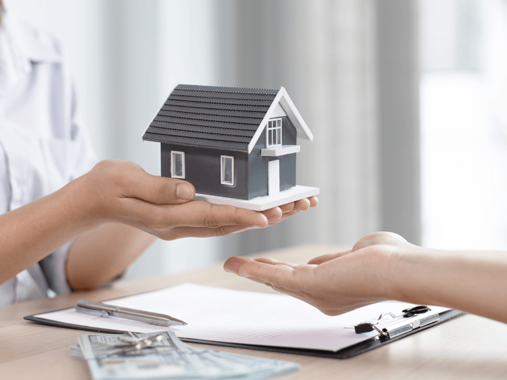 Modelo de casa sendo entregue entre duas pessoas sobre documentos, representando uma negociação de imóvel. Tudo para explicar quanto tempo demora para sair uma escritura de imóvel.