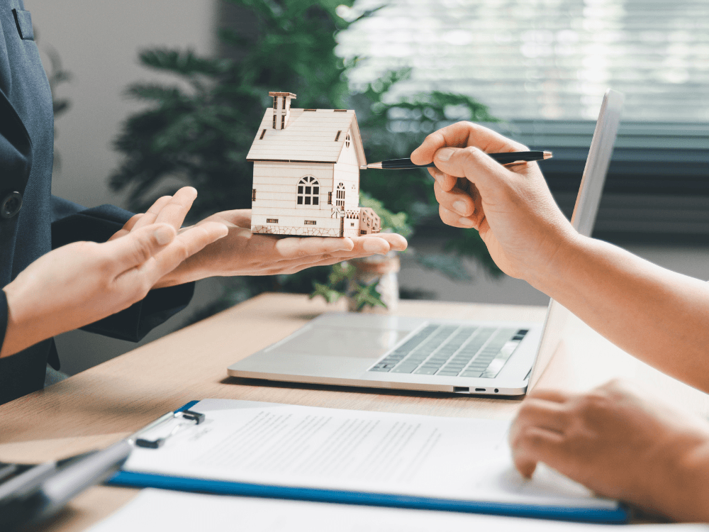 Pessoa apresenta uma maquete de casa enquanto outra aponta para o imóvel durante explicação sobre transcrição e matrícula imobiliária.