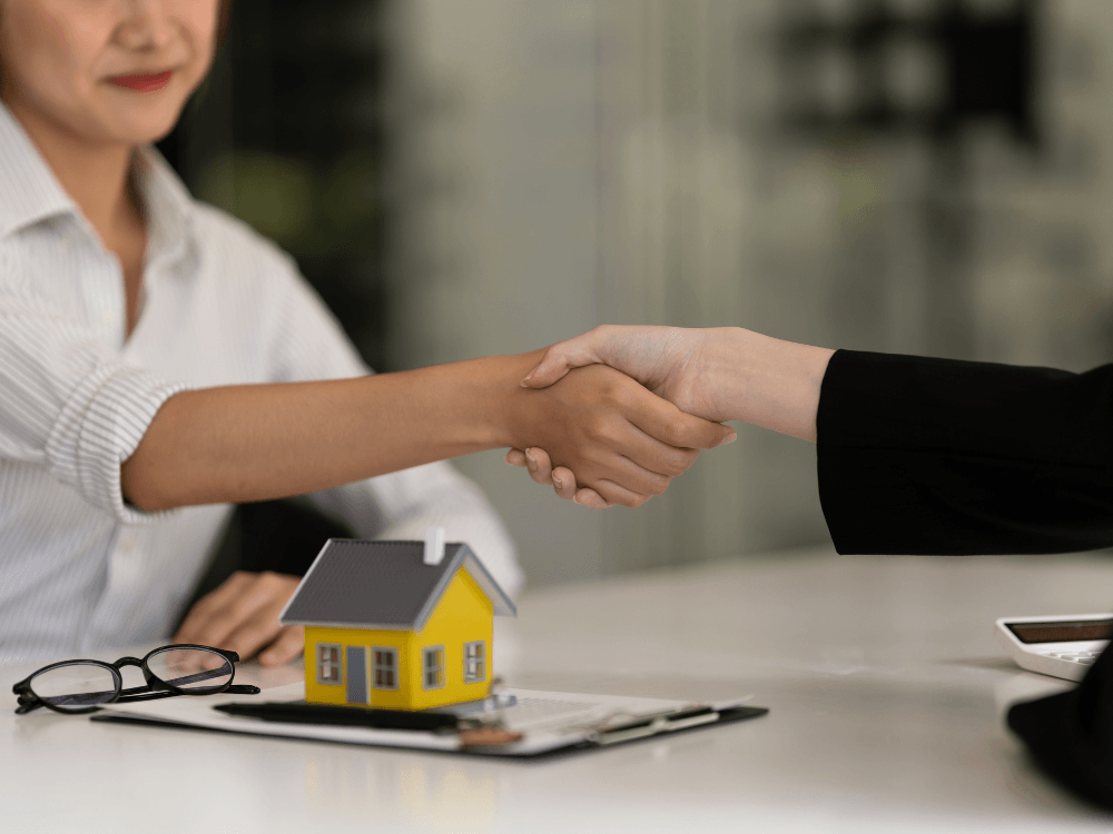 Duas pessoas apertam as mãos sobre uma mesa com maquete de casa e documentos, representando a conclusão da compra após o registro do imóvel.