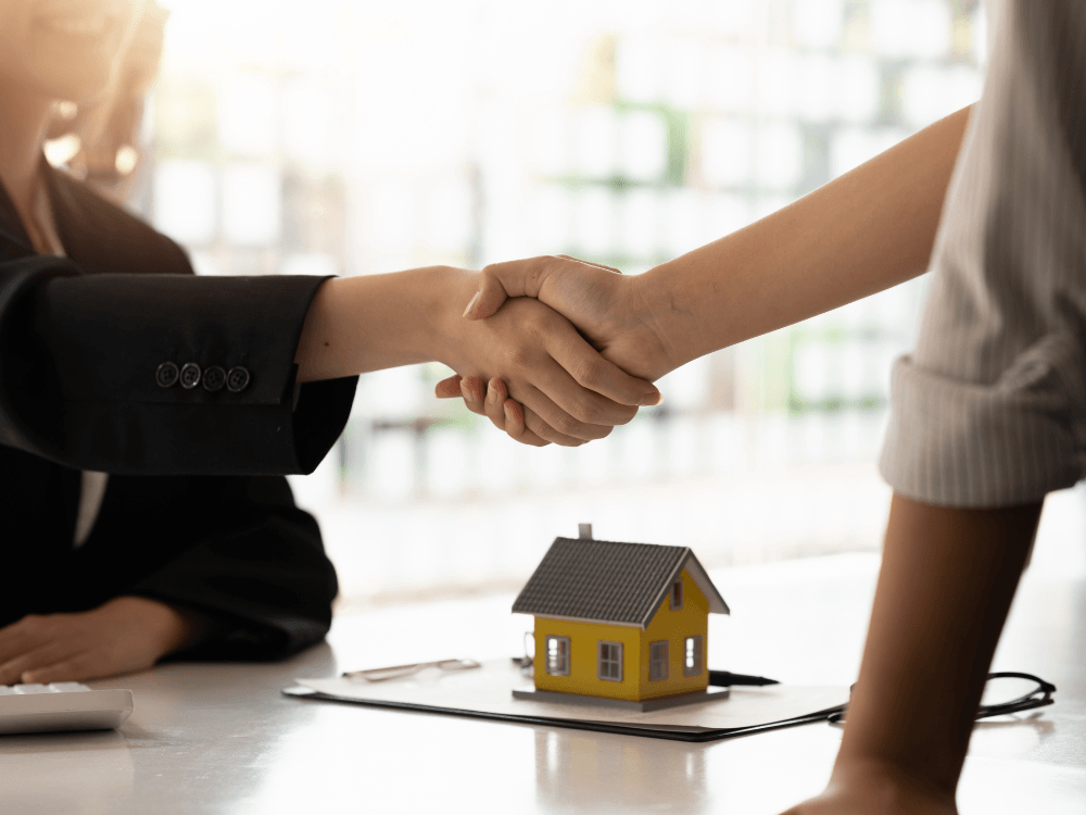 Duas pessoas apertam as mãos diante de uma maquete de casa, simbolizando um acordo para venda de imóvel com inquilino