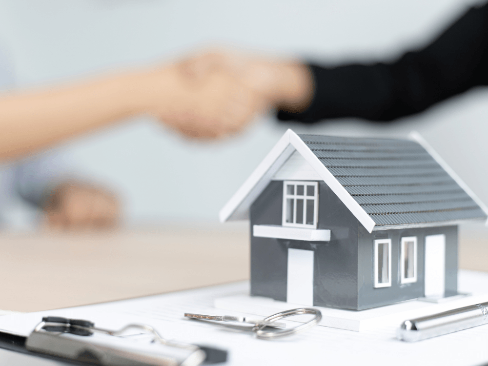 Maquete de uma casa sobre documentos enquanto duas pessoas apertam as mãos durante uma negociação imobiliária. Tudo para explicar as dicas para comprar apartamento na planta.