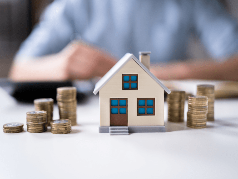 Casa em miniatura cercada por moedas representa os custos e valores envolvidos no aluguel de um imóvel residencial