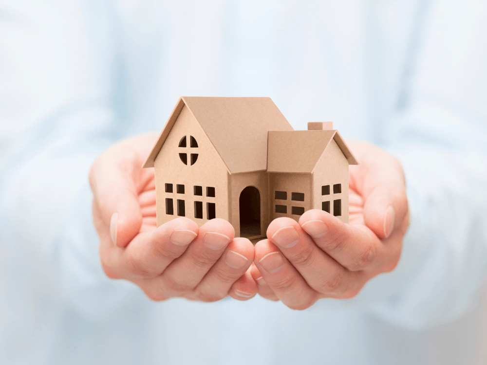 Modelo de casa em papelão apoiado nas mãos de uma pessoa, representando a escolha de um imóvel para comprar. Tudo para explicar como escolher um imóvel para comprar.