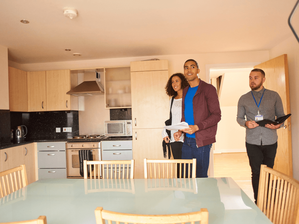 Casal visita cozinha de imóvel com armários de madeira enquanto corretor imobiliário apresenta detalhes da propriedade à venda.