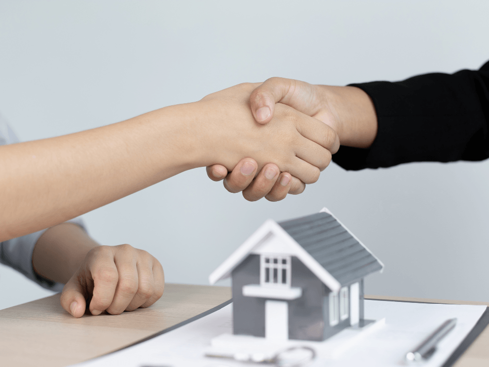 Duas pessoas apertam as mãos sobre uma mesa com maquete de casa e documentos, simbolizando negociação imobiliária.