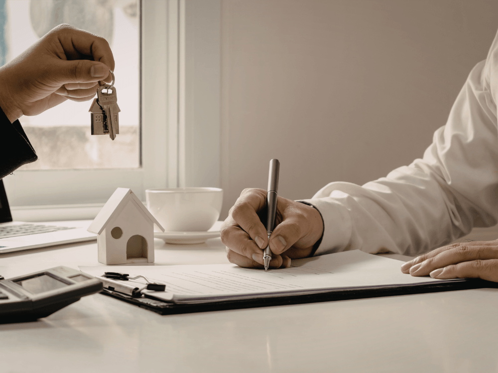 Pessoa assina documento enquanto outra segura chaves de uma casa, simbolizando uma negociação imobiliária formalizada.