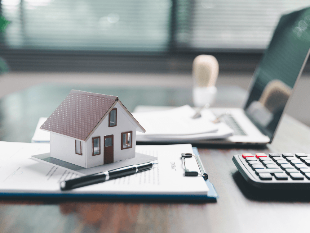 Maquete de uma casa sobre documentos imobiliários, com caneta e calculadora, representando a documentação de um imóvel. Tudo para explicar sobre os documentos de um imóvel