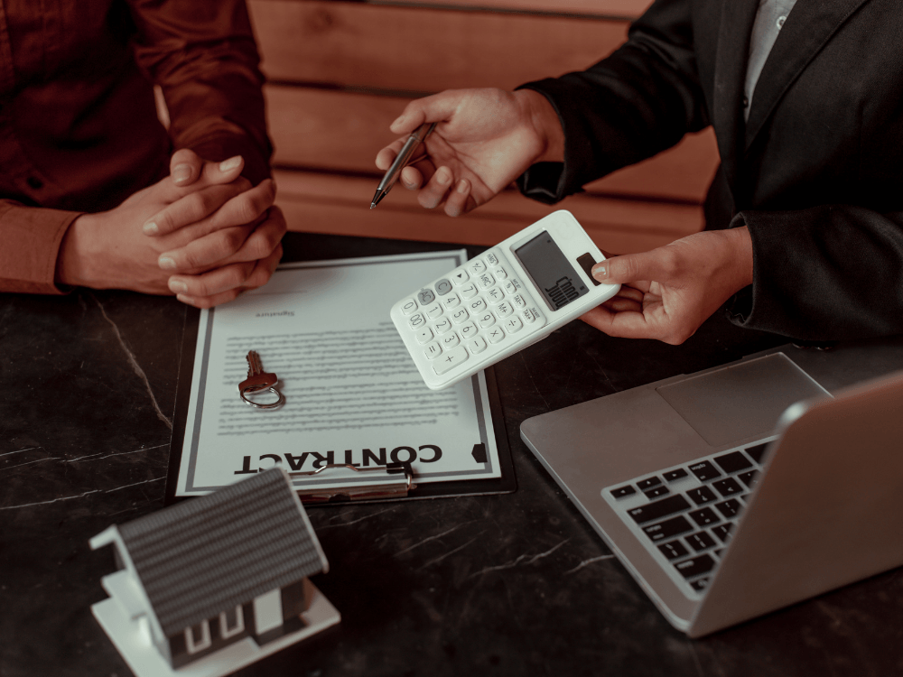 Contrato de imóvel sobre a mesa é analisado por duas pessoas, com calculadora, chaves e computador ao lado