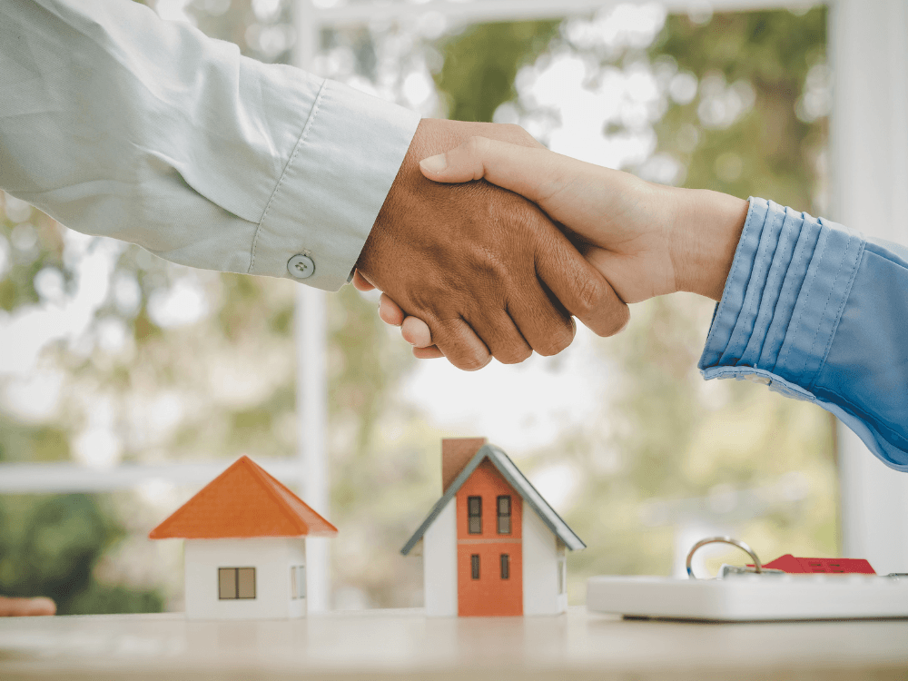 Negociação imobiliária representada por um aperto de mãos diante de miniaturas de casas e chaves sobre a mesa.
