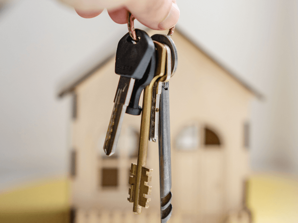 Chaves de uma casa em primeiro plano, com imóvel desfocado ao fundo, representando a compra de uma propriedade para investimento. Tudo para explicar se investir em imóveis é um bom negócio.