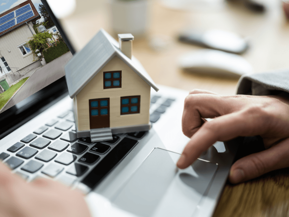 Miniatura de uma casa sobre um notebook, com uma mão no touchpad e uma fotografia de imóvel residencial ao fundo.