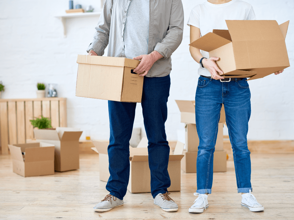 Casal transporta caixas de papelão em uma sala com outros volumes, mostrando a organização dos pertences durante a mudança.