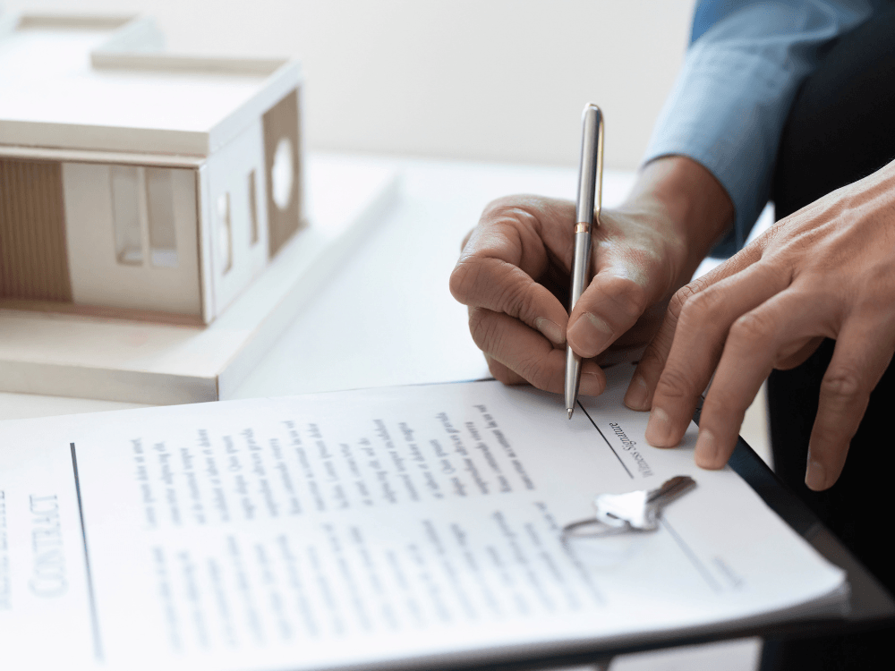 Pessoa assina um contrato imobiliário ao lado de uma maquete de casa e das chaves do imóvel sobre o documento.