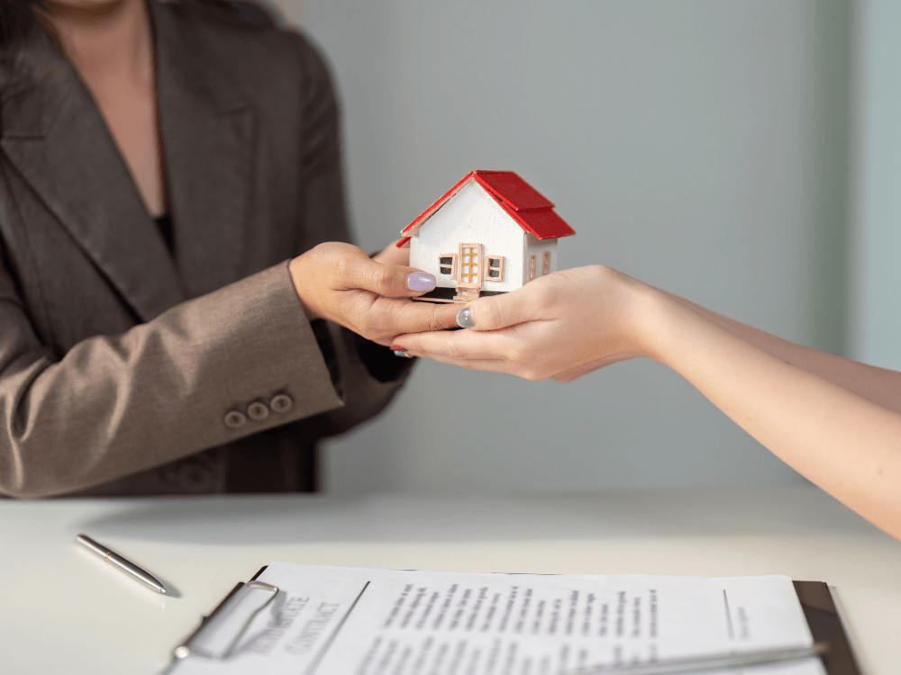 Duas pessoas seguram uma maquete de casa durante uma negociação imobiliária, simbolizando a transferência de propriedade.