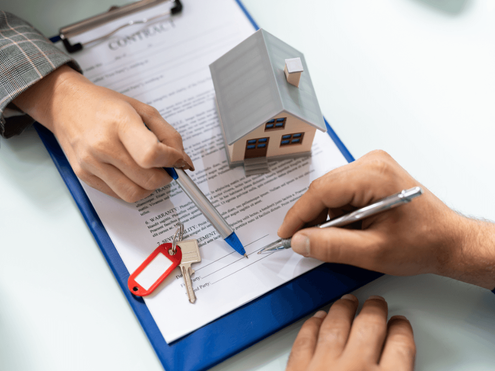 Mãos assinam contrato imobiliário sobre uma prancheta com maquete de casa e chaves, representando a entrega do imóvel novo.