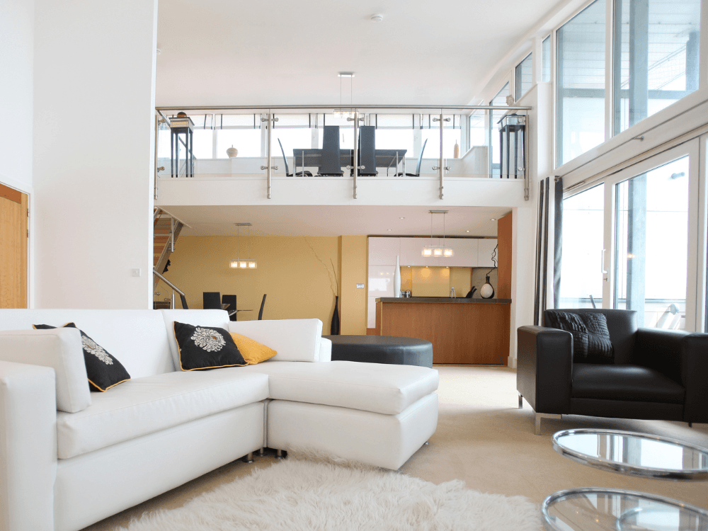 Sala ampla de apartamento duplex com sofá branco, cozinha ao fundo e mezanino com guarda-corpo de vidro. Tudo para explicar o que é apartamento duplex.