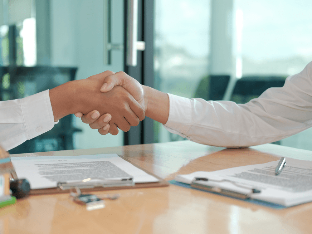Duas pessoas apertam as mãos sobre uma mesa com documentos e caneta, simbolizando um acordo relacionado à compra e venda de um imóvel.