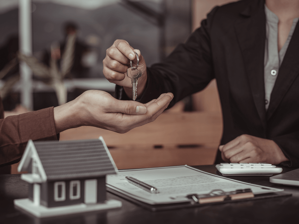 Profissional entrega um molho de chaves a outra pessoa sobre uma mesa com contrato imobiliário, calculadora e maquete de casa.