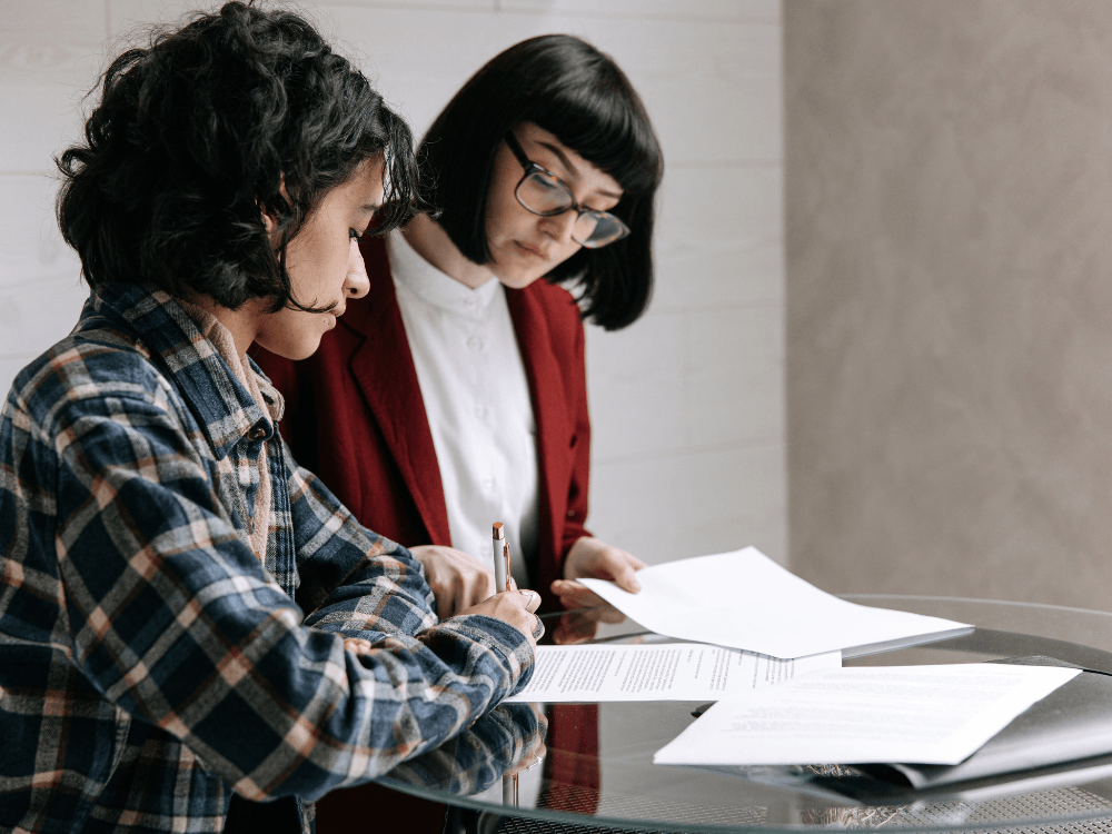 Mulher assina documentos enquanto outra profissional acompanha a leitura dos papéis durante uma análise documental relacionada a um imóvel. Tudo para explicar o que é ônus reais do imóvel.