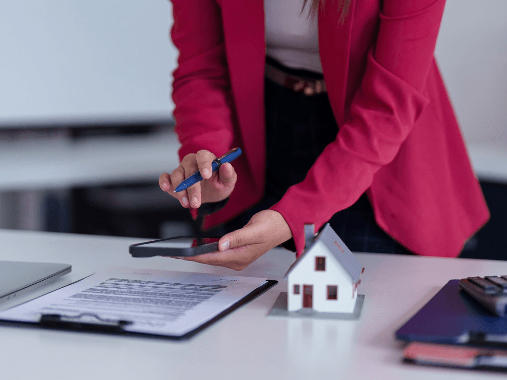 Profissional consulta documentos imobiliários com uma caneta e um celular ao lado de uma maquete de casa durante a análise de uma propriedade.