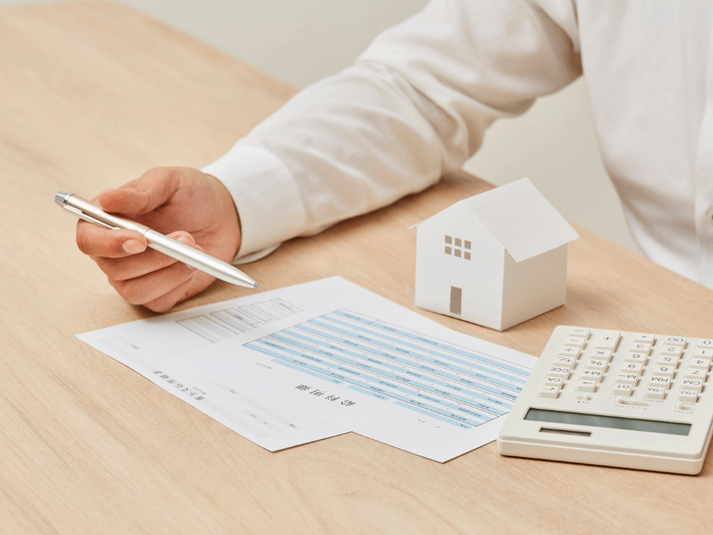 Mão segura uma caneta sobre um formulário financeiro ao lado de uma maquete de casa e uma calculadora, em análise imobiliária.